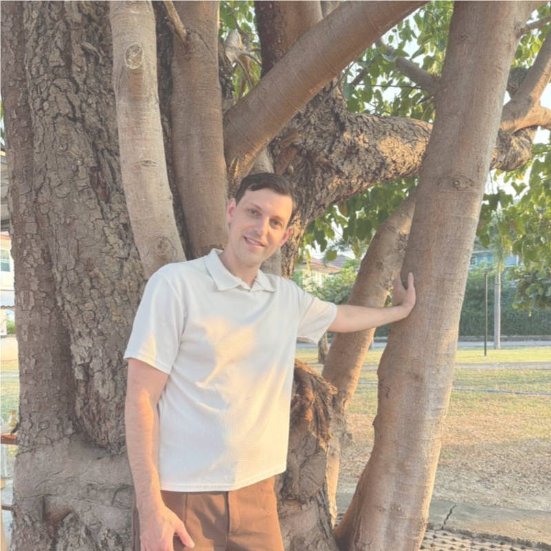 A man in a white polo shirt leaning against trees at sunset.